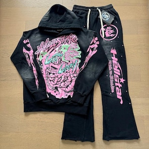 Könnte beinhalten: Schwarzes Hoodie- und passendes Hosen-Set. Der Hoodie hat eine rosa-grüne Grafik mit dem Text "HELLSTAR N GUTS GLORY!" und rosa Armdesigns. Die Hose hat einen Kordelzug in der Taille und das Wort "Hellstar" in Rosa.
