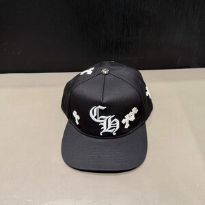 Può includere: Un cappellino da baseball nero con un logo CH ricamato in bianco in stile gotico sul pannello frontale, accentuato da motivi a croce bianchi sui lati e un piccolo bottone metallico argentato al centro della parte superiore.