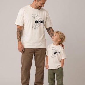 Puede incluir: Dos camisetas de manga corta en color blanco roto con estampados gráficos en negro. Una camiseta muestra el texto Ctrl+C con un patrón geométrico, mientras que la otra muestra el texto Ctrl+V con un patrón a juego, ambas en un estilo sencillo.