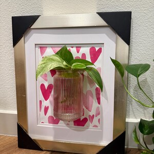 Puede incluir: Una obra de arte enmarcada con un jarrón de vidrio que contiene una planta. El fondo presenta corazones rosas. El marco tiene un acabado dorado y esquinas negras. Una pieza decorativa.