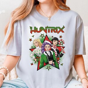 May include: A light gray t-shirt featuring a graphic of three women in a Christmas-themed design. The word "HUNTRIX" is displayed above the graphic. The women are surrounded by stars and Christmas decorations.