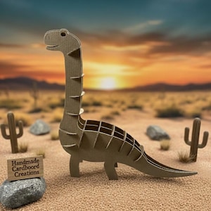 Könnte beinhalten: Ein Papp-Dinosaurier-Modell mit langem Hals und Schwanz steht in einer Wüstenlandschaft mit Kakteen und einem Sonnenuntergang im Hintergrund. Der Dinosaurier besteht aus Wellpappe. Ein kleines Holzschild trägt die Aufschrift "Handmade Cardboard Creations."