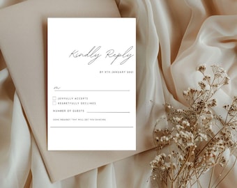 May include: A white wedding RSVP card with the words "Kindly Reply" in elegant script. The card includes checkboxes for accepting or declining, a space for the number of guests, and a line for a song request. The card is on a beige envelope.