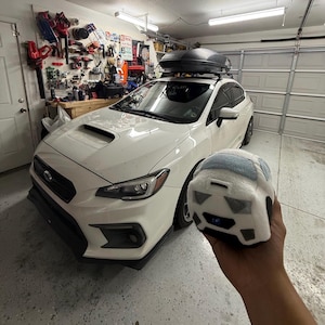 May include: A white Subaru sedan with a black roof cargo carrier parked in a garage. A plush toy car in the shape of a Subaru is held in front of the car. The garage has tools and equipment on the walls.