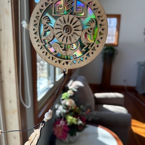 May include: Circular wooden suncatcher with a holographic centre. The design includes a sun, geometric shapes, and two figures. The outer edge has a repeating triangle pattern. A wooden feather and beads hang below the suncatcher.