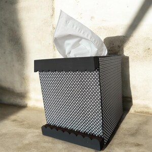 Könnte beinhalten: Ein Taschentuchspender mit schwarz-weißem Muster und gewelltem Rand. Die Box hat ein geometrisches Muster und ein weißes Taschentuch wird von oben herausgezogen. Der Spender besteht aus einem robusten Material.