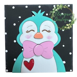 May include: A wooden penguin decoration with a teal head and body, a pink bow tie, and a red heart. The penguin is set against a black background with white polka dots. The text "Unique Boutique" is in a gold circle.