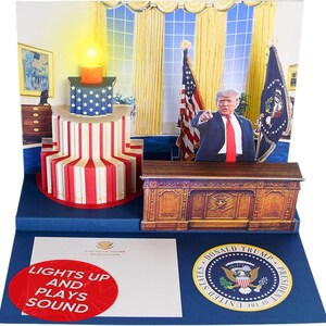 May include: A pop-up greeting card featuring a 3D White House Oval Office scene with a red, white, and blue cake, a figure of Donald Trump, and American flags. The base includes the Presidential Seal and text reading LIGHTS UP AND PLAYS SOUND.