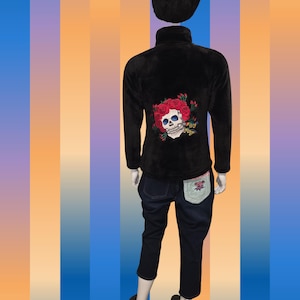 May include: Black fleece zip-up jacket with a skull and rose embroidery on the back. The outfit includes dark wash denim capri pants and a black feathered headpiece. The back pocket of the jeans has a floral design.