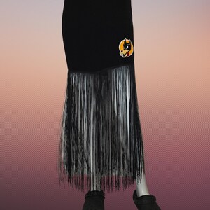 May include: Black skirt with long fringe and a cartoon dog patch. The skirt has a straight silhouette and is made of a solid black material. The fringe is black and white. The patch features a cartoon dog with a yellow background.