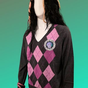 Könnte beinhalten: Ein dunkelgrauer V-Ausschnitt-Pullover mit einem rosa-lila Rautenmuster. Der Pullover hat einen runden Aufnäher auf der Brust und wird auf einer Schaufensterpuppe präsentiert. Die Schaufensterpuppe trägt eine Perücke mit schwarzen Haaren und rosa und blonden Strähnen.