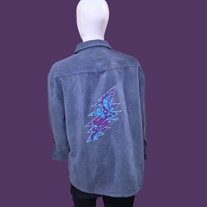 May include: A gray button-down shirt with a lightning bolt design on the back. The lightning bolt is a combination of blue and purple. The shirt is displayed on a mannequin against a purple background.