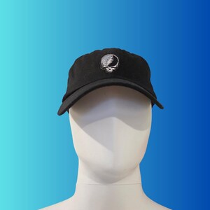 May include: Black baseball cap with a curved brim. The cap features a white and gray emblem on the front. The hat is displayed on a mannequin against a blue gradient background. The cap is a classic style.