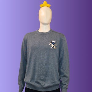 Puede incluir: Un suéter gris de cuello redondo con un pequeño gráfico de oso en el pecho. El maniquí lleva un sombrero amarillo. El suéter se exhibe en un maniquí sobre un fondo degradado púrpura.