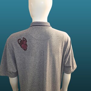 May include: Grey polo shirt with a dark purple collar and a heart-shaped embroidered design on the back. The shirt is displayed on a mannequin against a teal background. The heart design has red and purple accents.