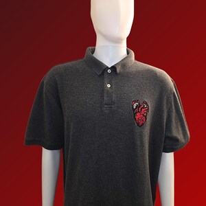 May include: Dark grey polo shirt with short sleeves and a three-button placket. A red and black embroidered patch is on the left chest. The shirt is displayed on a white mannequin against a red background.