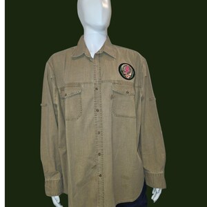 May include: A long-sleeved, button-down shirt in a khaki color, featuring two chest pockets and a patch on the upper left. The patch has a circular design with floral and text elements. The shirt is displayed on a mannequin.