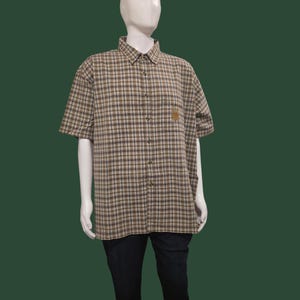 May include: A short-sleeved, button-down plaid shirt in shades of brown, tan, and black, displayed on a mannequin. The shirt has a small embroidered emblem on the chest and is paired with dark blue capri pants. The mannequin is set against a dark green backdrop.