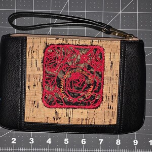 Könnte beinhalten: Schwarze und Kork-Handgelenktasche mit rotem Blumendesign. Die Handgelenktasche hat einen schwarzen Griff und einen goldenen Reißverschluss. Das Mittelteil ist aus Kork mit einem roten, gestickten Blumendesign. Die Seiten sind schwarz.