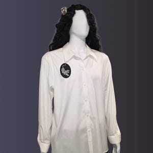 May include: A white button-down shirt with long sleeves and a black oval patch featuring a detailed leopard design. The shirt is displayed on a mannequin with dark hair and a decorative hair accessory.