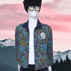 May include: A mannequin wearing a colorful patterned jacket with a blue collar and a small patch. The jacket has a button-up front and is paired with dark blue jeans. The mannequin also wears a black wig and yellow-rimmed glasses.