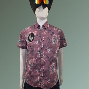 May include: A mannequin wearing a short-sleeved button-down shirt with a floral pattern in shades of purple, blue, and white. The shirt has a small patch on the chest and is paired with dark blue jeans. The mannequin also wears a black wig with rainbow-tinted goggles.