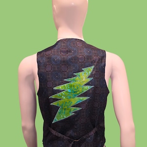 May include: A black vest with a patterned design and a green and blue lightning bolt applique. The vest is displayed on a mannequin against a green background. The vest has a tie in the back.
