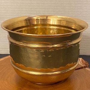 May include: A polished brass planter with a wide, flared rim and a rounded body. The central section of the vessel features a distinct hammered texture, while the upper and lower portions remain smooth, creating a contrast in surface finish.