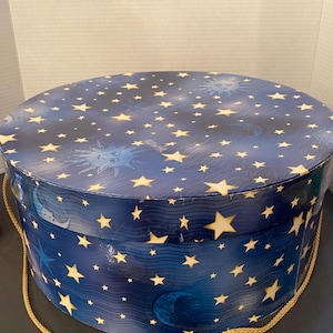 May include: A round, lidded storage box featuring a deep blue background with a pattern of yellow stars, crescent moons, and sun faces. The box has a smooth, glossy finish and includes a gold-coloured braided rope handle attached to the sides.