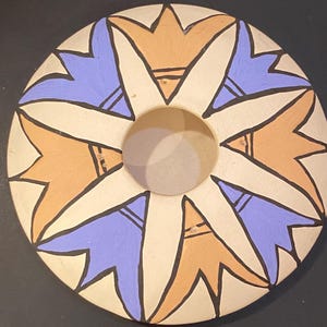 May include: A circular ceramic vessel with a central opening, featuring a repeating star-like pattern in alternating light purple and muted orange petals outlined in black against a cream-colored background, creating a symmetrical geometric design.