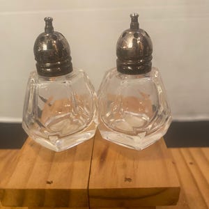 Op de afbeelding: Een paar zout- en peperstrooiers van helder, gefacetteerd glas met metalen, koepelvormige doppen voorzien van kleine gaatjes. De strooiers hebben een geometrisch ontwerp met meerdere zijden en zijn leeg van binnen.
