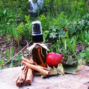 May include: A brown glass bottle with a black spray top and a natural fiber bow. The bottle is sitting on a tree stump with cinnamon sticks and a red apple.