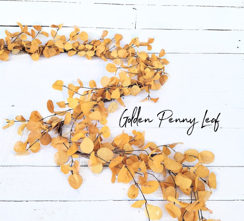 May include: A garland of golden penny leaf eucalyptus branches on a white wood background. The text "Golden Penny Leaf" is written in black cursive.