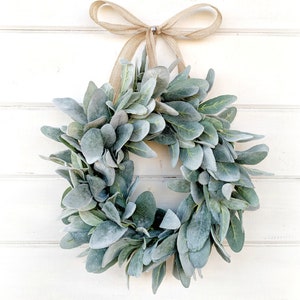 May include: A green and gray faux lamb's ear wreath with a burlap bow hanging on a white wooden wall.