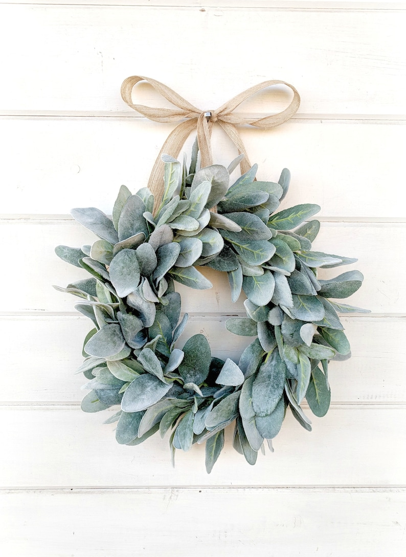 May include: A green and gray faux lamb's ear wreath with a burlap bow hanging on a white wooden wall.