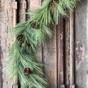 May include: A green pine garland with brown pine cones draped over a rustic wooden door.