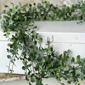 May include: A green leafy garland with small leaves, draped over a white vintage suitcase. The garland is made of artificial greenery and is perfect for adding a touch of nature to any space.