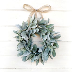 May include: A green and gray lamb's ear wreath with a brown burlap ribbon bow hanging on a white wooden wall.