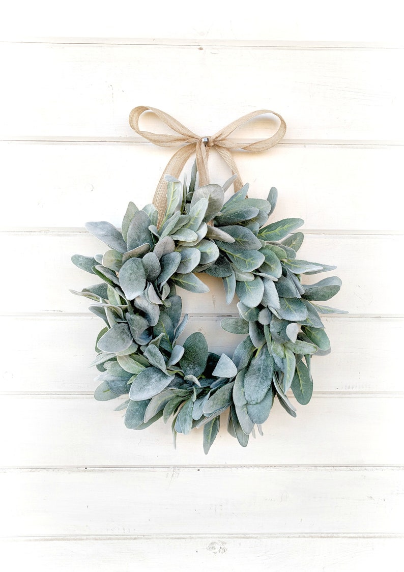 May include: A green and gray lamb's ear wreath with a brown burlap ribbon bow hanging on a white wooden wall.