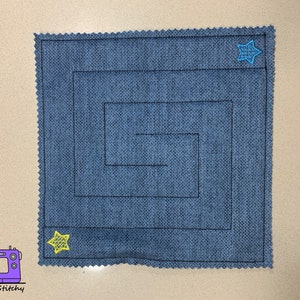 May include: A blue fabric square with a black stitched maze pattern and two yellow and blue star shapes.