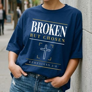 May include: A navy blue t-shirt featuring the words "BROKEN BUT CHOSEN" in white and gold. A white cross graphic is below the text, with the Bible verse Ephesians 2:8 printed underneath. The t-shirt is a relaxed fit.
