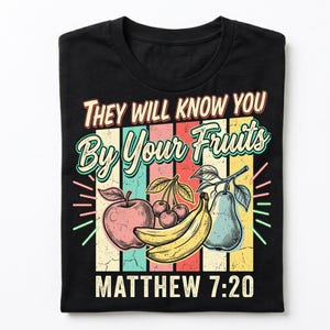 May include: Black t-shirt with a colorful graphic featuring an apple, cherries, a banana, and a pear. The text reads "THEY WILL KNOW YOU BY YOUR FRUITS" and "MATTHEW 7:20" in a vintage style.