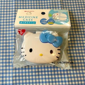 Puede incluir: Un estuche para medicamentos de Hello Kitty en un embalaje de plástico transparente. El estuche blanco tiene la forma de la cara de Hello Kitty, con un lazo azul, ojos negros y una nariz amarilla. El embalaje incluye texto en inglés y japonés.