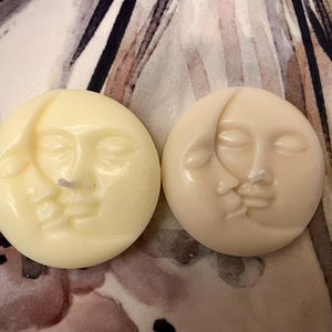 Puede incluir: Dos velas redondas de color blanquecino con un diseño de luna creciente y rostro. Una vela es de un tono más claro que la otra. Las velas tienen una superficie lisa y una pequeña mecha. El fondo es un estampado floral.