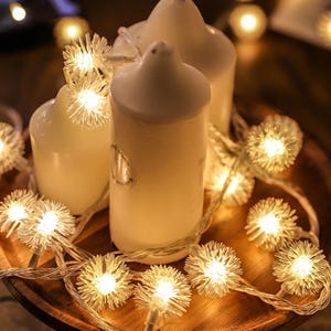 May include: A close-up of three white pillar candles with a string of small, glowing, starburst-shaped lights. The candles are arranged on a wooden surface, creating a warm and inviting ambiance. The lights add a festive touch.