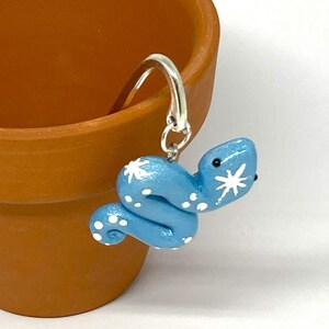 May include: A light blue snake earring with white star and dot details hangs near a terracotta pot. The earring is made of polymer clay and has a silver-colored hook. The pot is empty and has a round shape.
