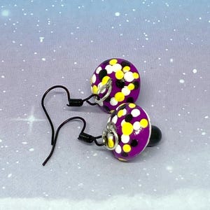 以下が含まれることがあります： 黒いフックが付いた紫色のイヤリングのペア。丸いイヤリングは、黄色と白のドットのパターンが特徴です。イヤリングは、青と白の空の背景に配置されています。