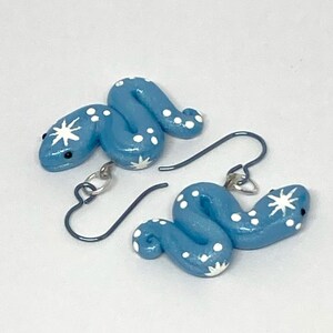 May include: A pair of light blue snake-shaped earrings. Each earring has a coiled design with white polka dots and a starburst pattern on the head. The earrings have silver-coloured hooks.
