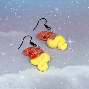 May include: A pair of novelty earrings featuring a red and yellow design. The earrings have a red, textured top section and a yellow, fluffy bottom section. The earrings hang from black ear wires.