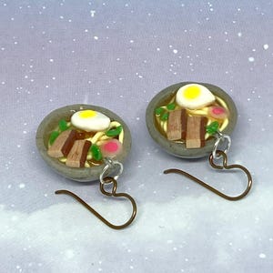 May include: A pair of miniature ramen bowl earrings. Each earring features a gray bowl filled with noodles, a fried egg, and other toppings. The earrings have bronze-colored hooks and are set against a blue and white speckled background.